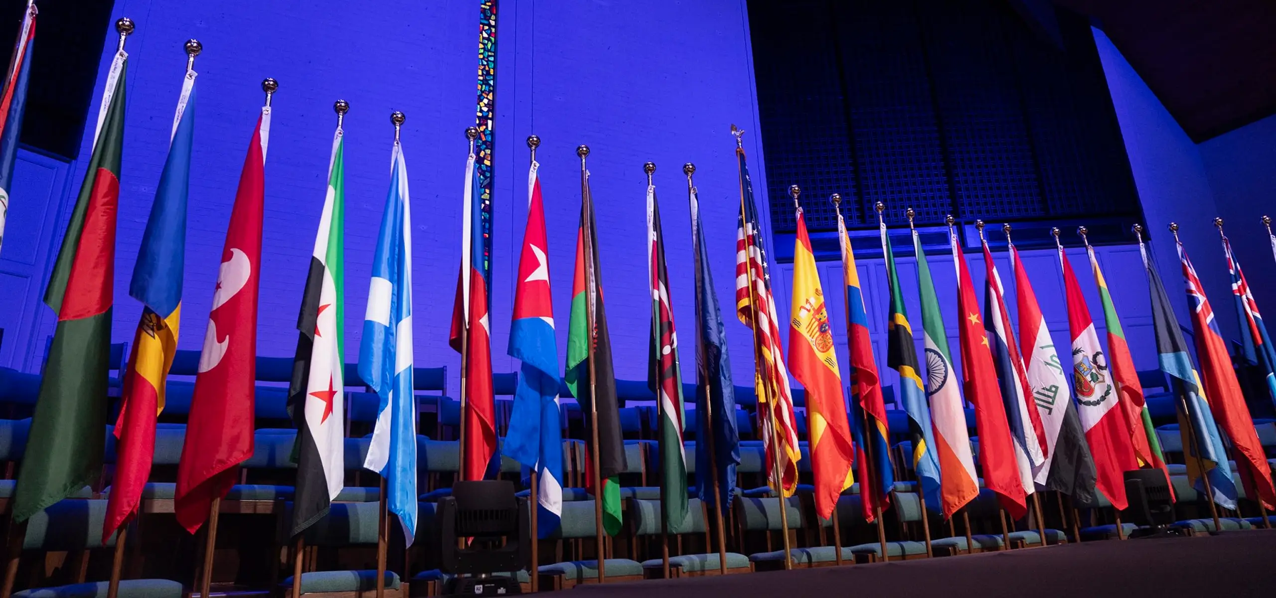 Row of various international flags displayed inside a church sanctuary with blue lighting and seating in the background.