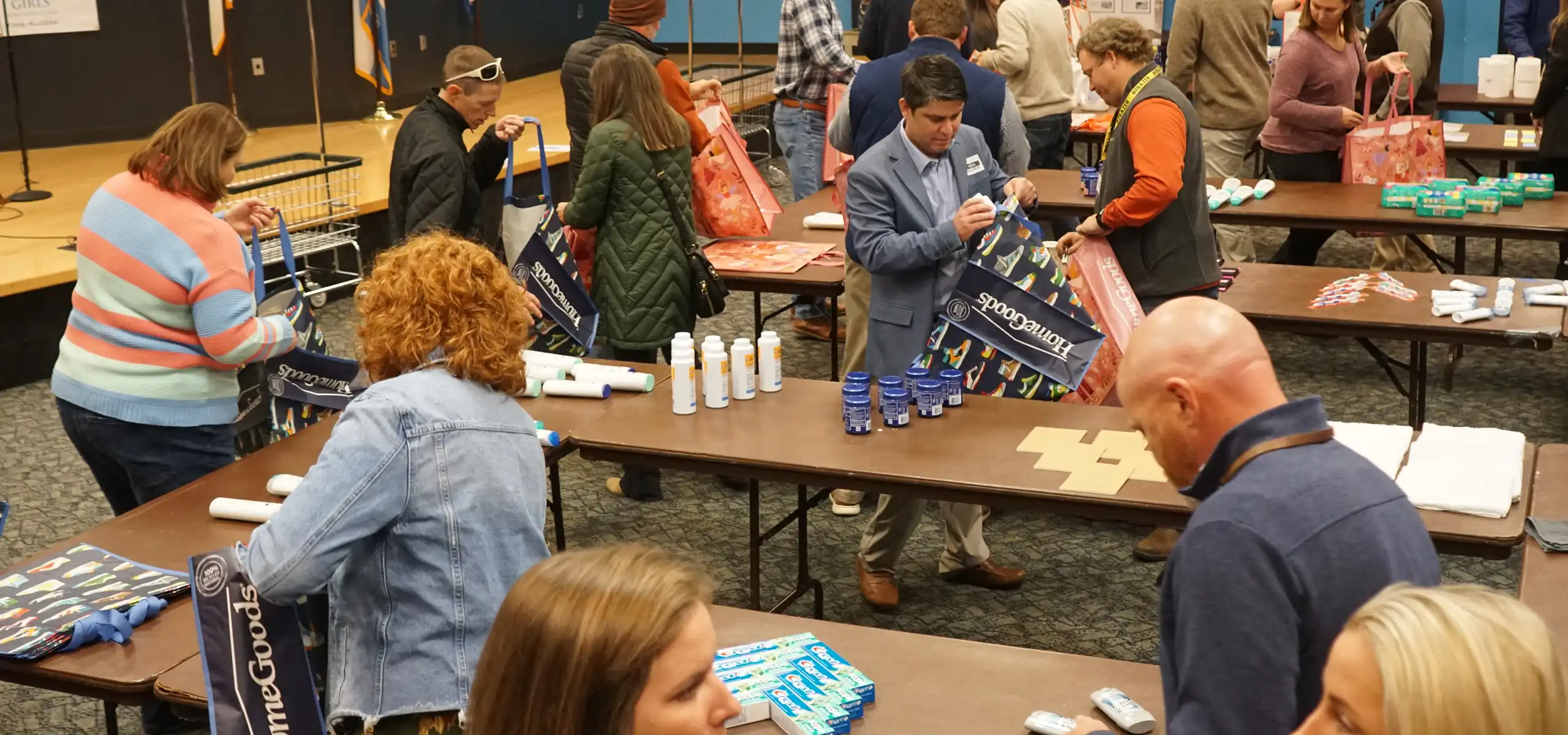 People standing around tables packing various toiletry items like toothpaste, lotion, and ointments into reusable bags at an indoor event.