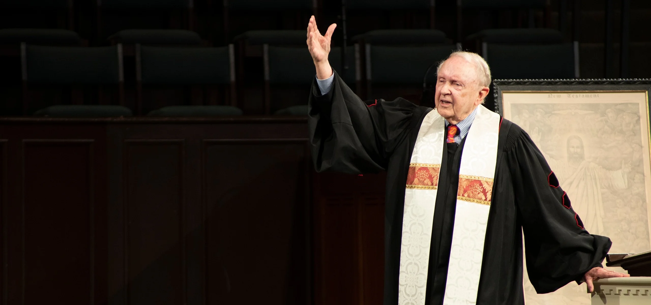 Elderly pastor in black robe and white stole preaching with raised hand inside church.