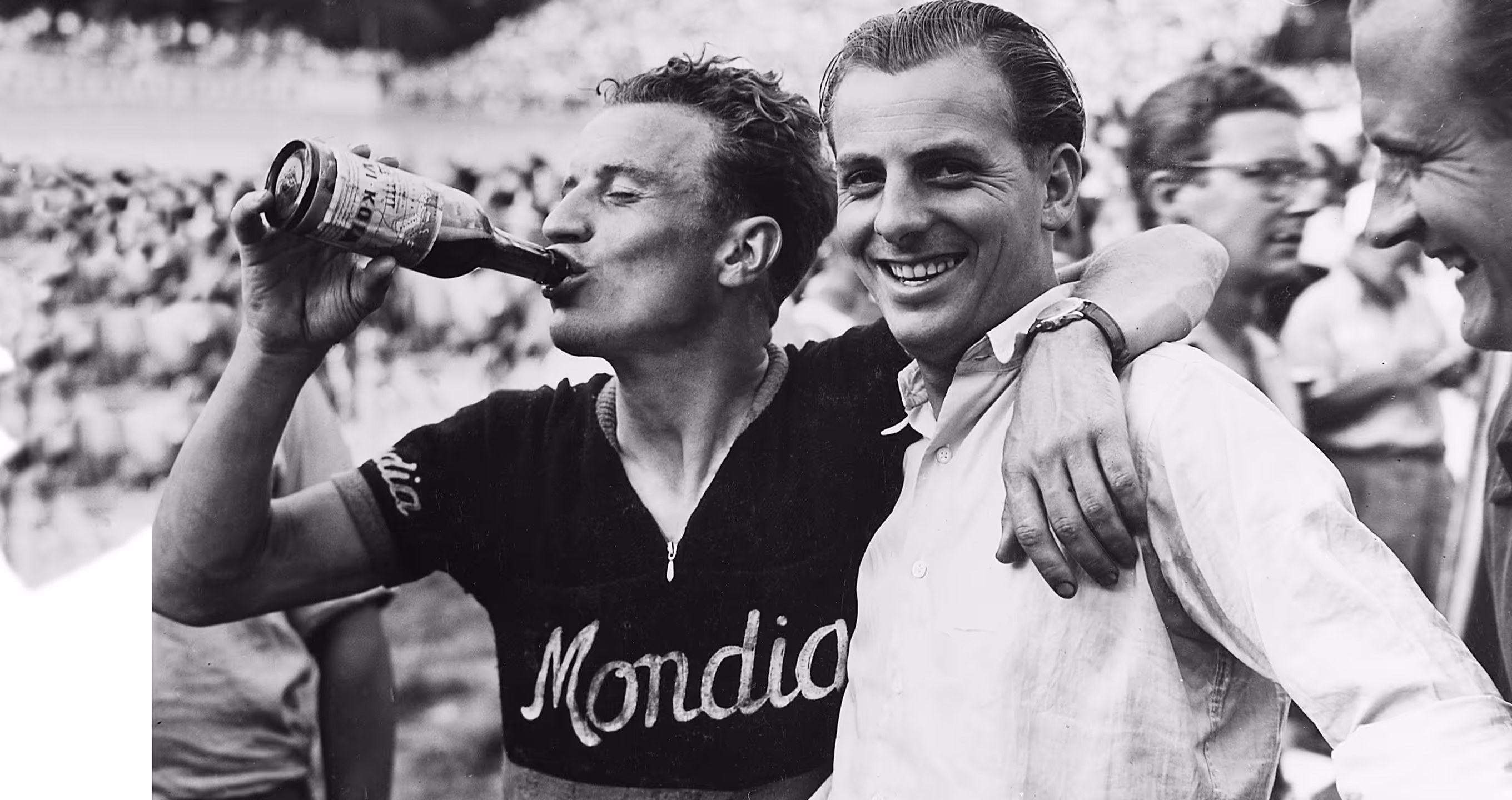 A cyclist wearing a Mondia jersey drinks from a bottle while smiling with two men, one with his arm around the cyclist's shoulders.