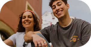 Two smiling young people, a woman and a man, sitting outdoors with blurred background buildings.