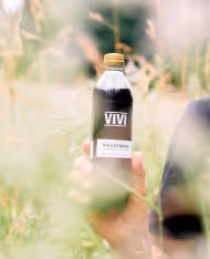 Hand holding a VIVI bottled drink labeled 'Kola Original' outdoors with green foliage in the background.