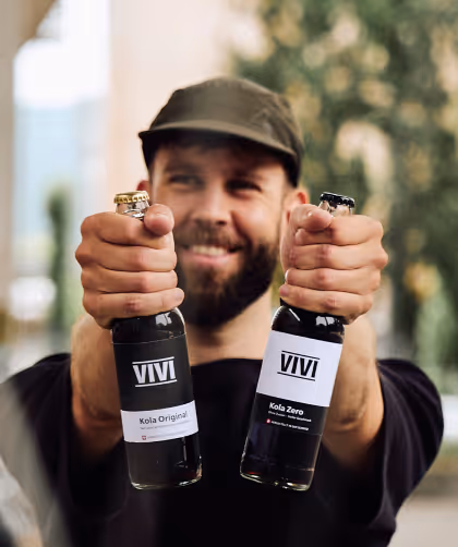 Smiling man wearing a cap holding two VIVI brand cola bottles labeled Kola Original and Kola Zero.