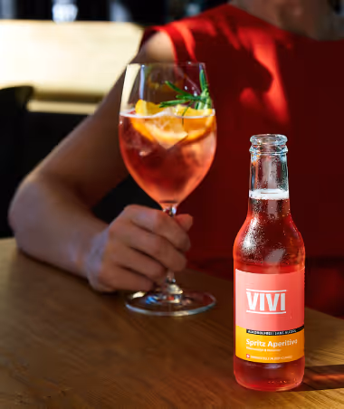 Person in red holding a wine glass with an orange spritz cocktail garnished with herbs, next to a chilled VIVI Spritz Aperitivo bottle on a wooden table.