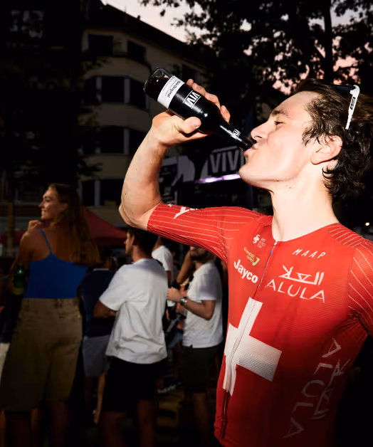 Cyclist in red Swiss team uniform drinking from a bottle at an outdoor event during dusk.