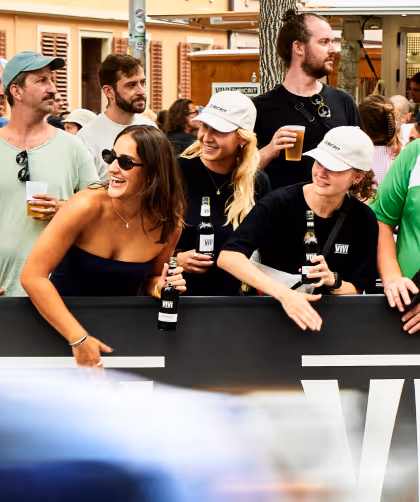Group of people smiling and holding drinks at an outdoor event behind a black barrier.