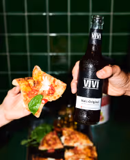 Hand holding a bottle of VIVI Kali Original beer and another hand holding a slice of pizza with basil leaf.