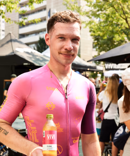 Man in a pink cycling jersey holding a bottle of VIVI iced tea at an outdoor event with people and tents in the background.