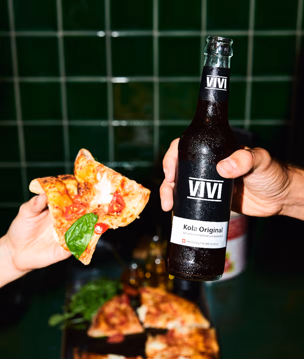 Eine Hand hält eine Pizzascheibe mit Basilikum, eine andere eine Flasche VIVI Kola Original vor grün gekacheltem Hintergrund.