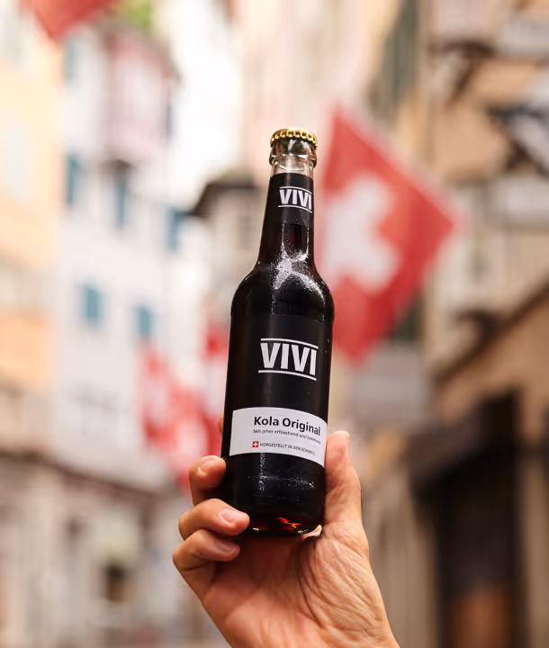 Hand hält eine Flasche Vivi Kola Original vor unscharfem Hintergrund mit Schweizer Fahnen.