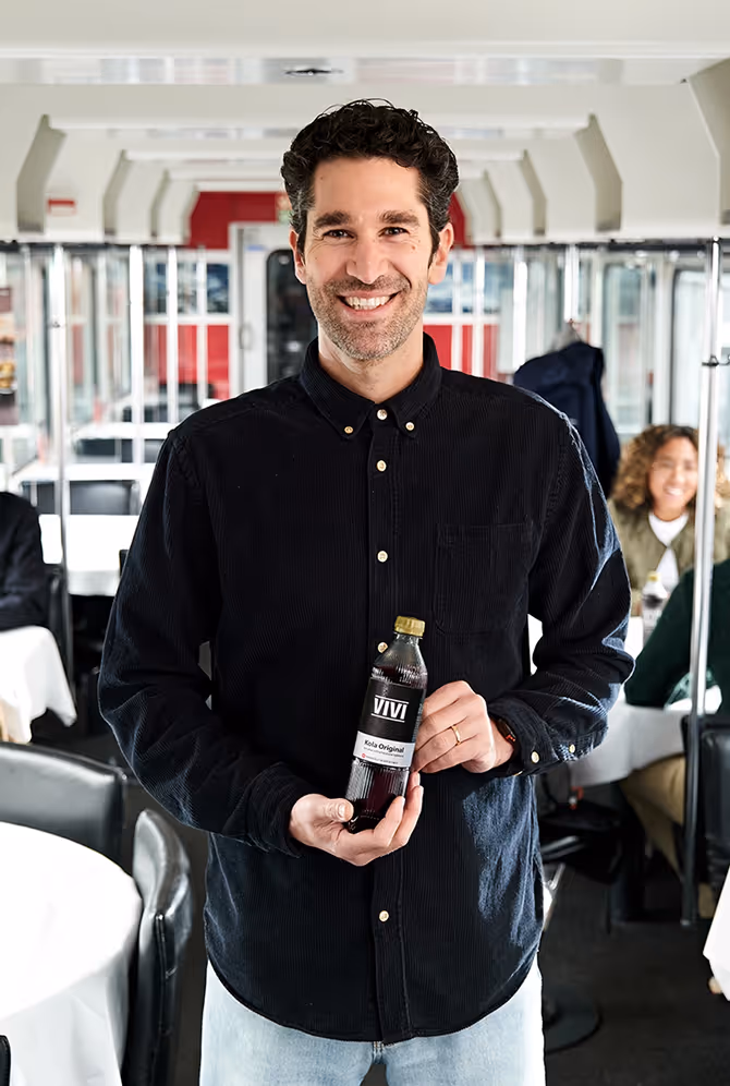 Ein lächelnder Mann in schwarzem Hemd hält eine Flasche VIVI Kola Original in einem Restaurant.