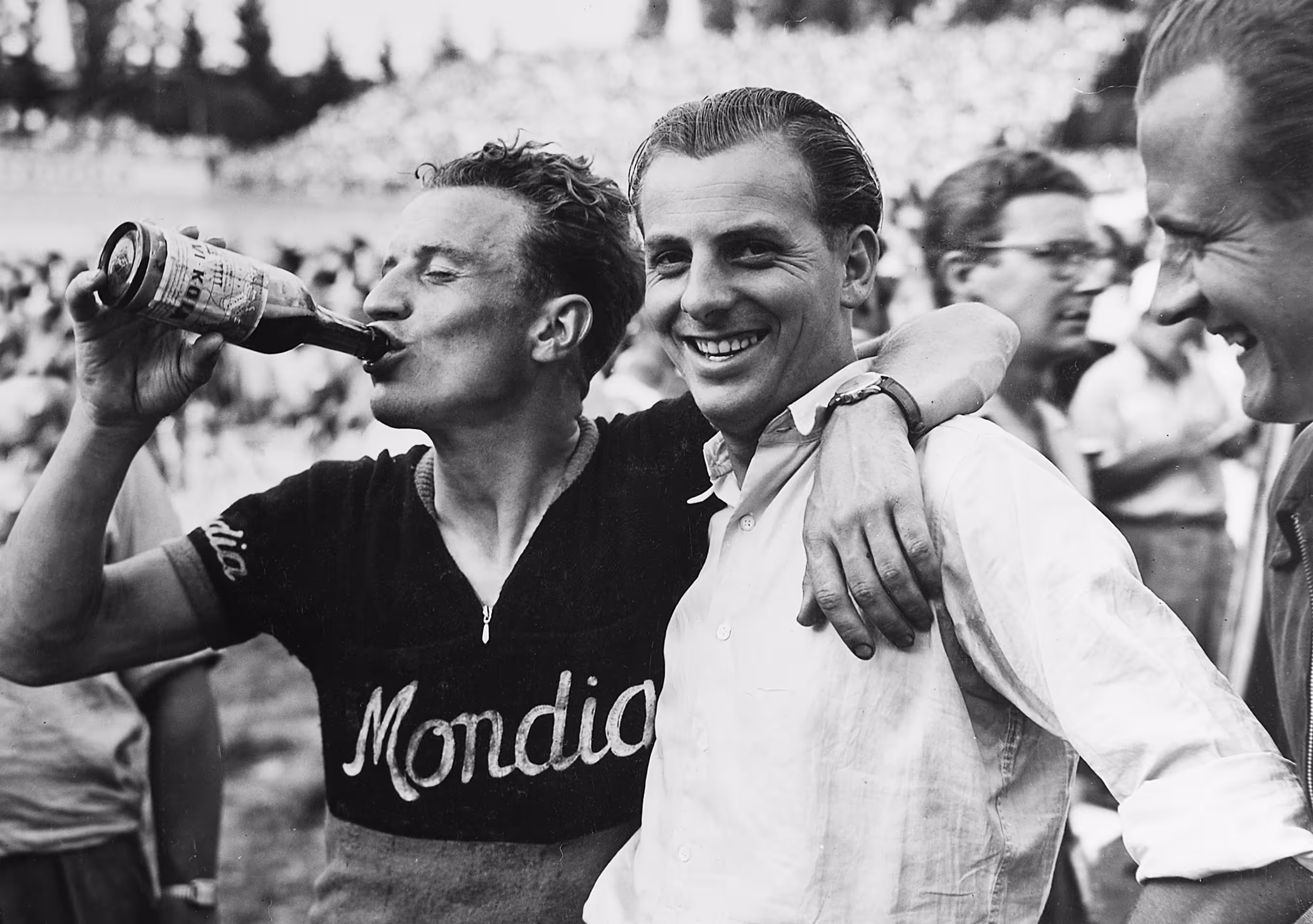 A cyclist wearing a Mondia jersey drinks from a bottle while smiling with two men, one with his arm around the cyclist's shoulders.