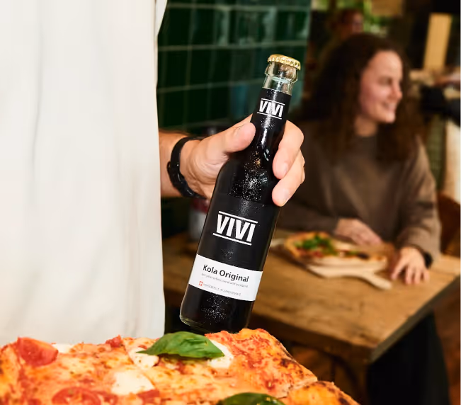 Hand hält eine Flasche VIVI Kola Original neben einem Teller mit Pizza, im Hintergrund eine lächelnde Frau am Tisch.