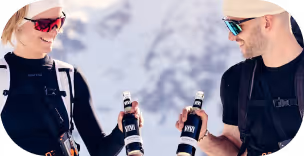 Zwei Personen in Skikleidung und Sonnenbrillen stoßen mit Flaschen an vor einer verschneiten Berglandschaft.