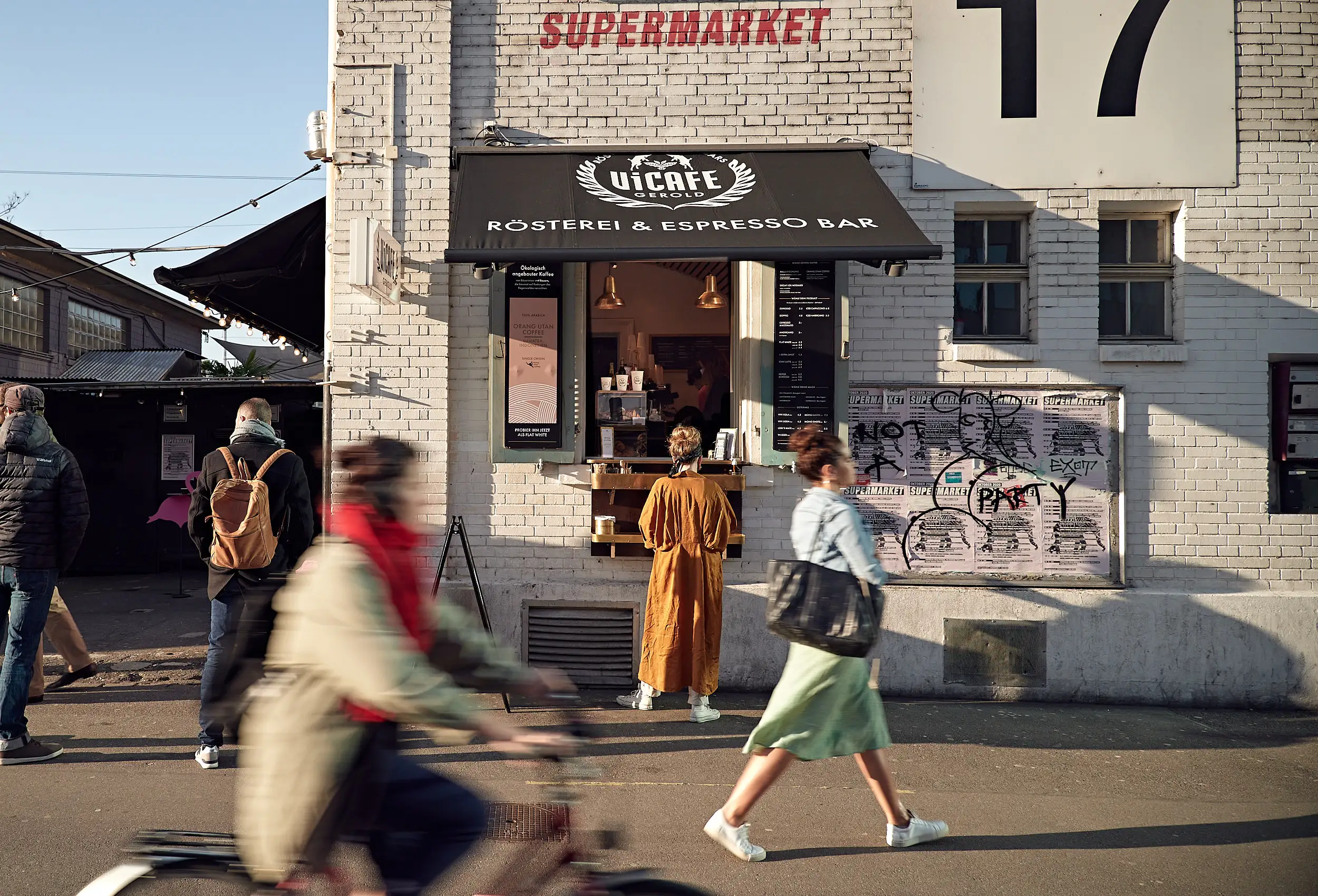 Person in braunem Mantel steht vor einem Fenster einer Kaffeebar namens ViCafe Gerold mit schwarzem Sonnenschutz und der Aufschrift Rösterei & Espresso Bar.