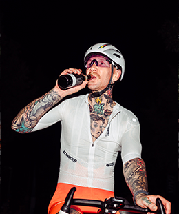 Bearded man in a cap drinking from a glass bottle outdoors with urban buildings in the background.