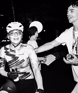 Smiling female cyclist in black and white VIVI Zurich kit holding water bottles with others' hands reaching in front of a VIVI Zurich backdrop.