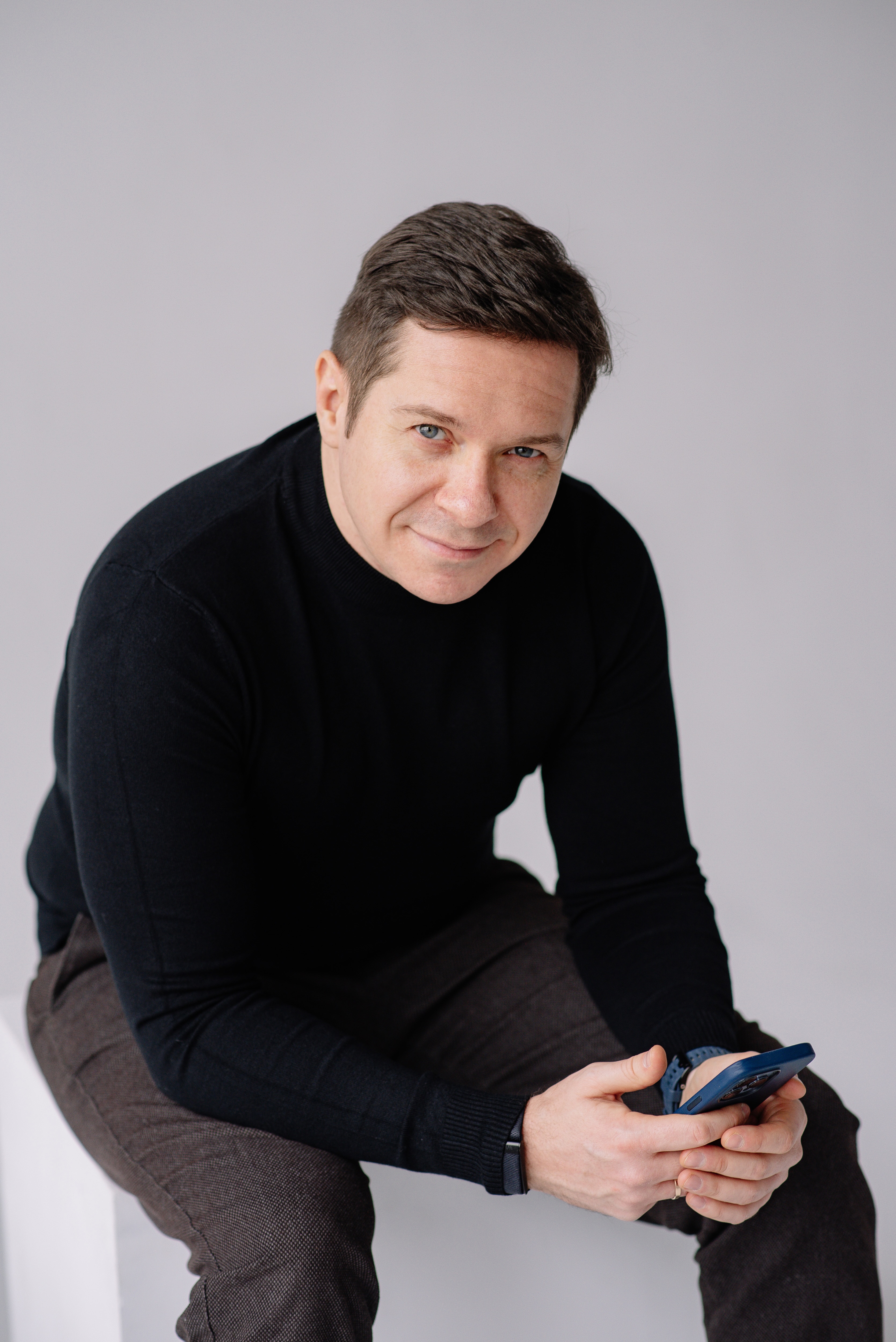Smiling man wearing a black sweater and brown pants, sitting and holding a smartphone.