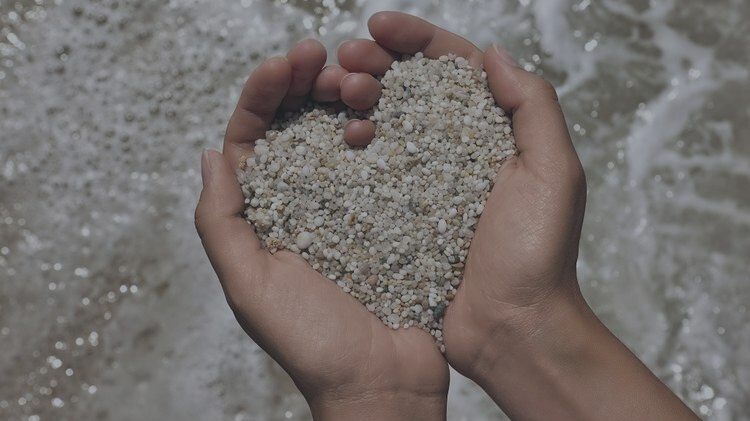 Mains formant un cœur rempli de sable face à la mer, symbole de douceur, intention et connexion.