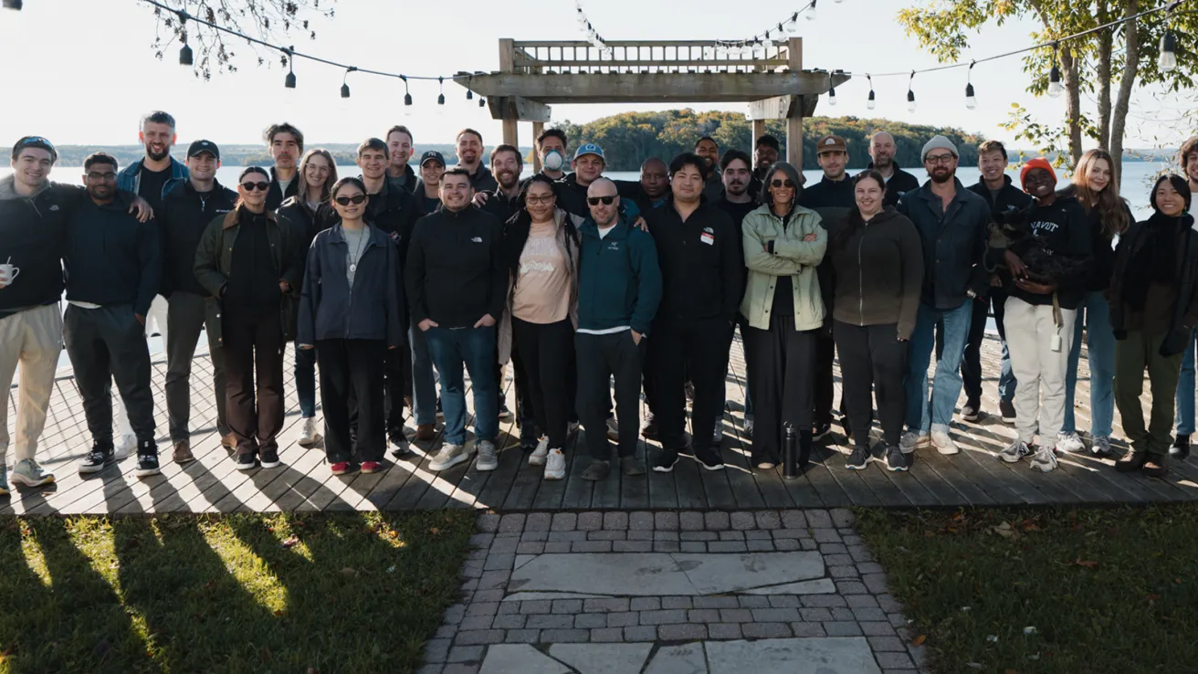 Group of diverse people standing together on a wooden deck with a pergola and string lights, overlooking a lake in the background.