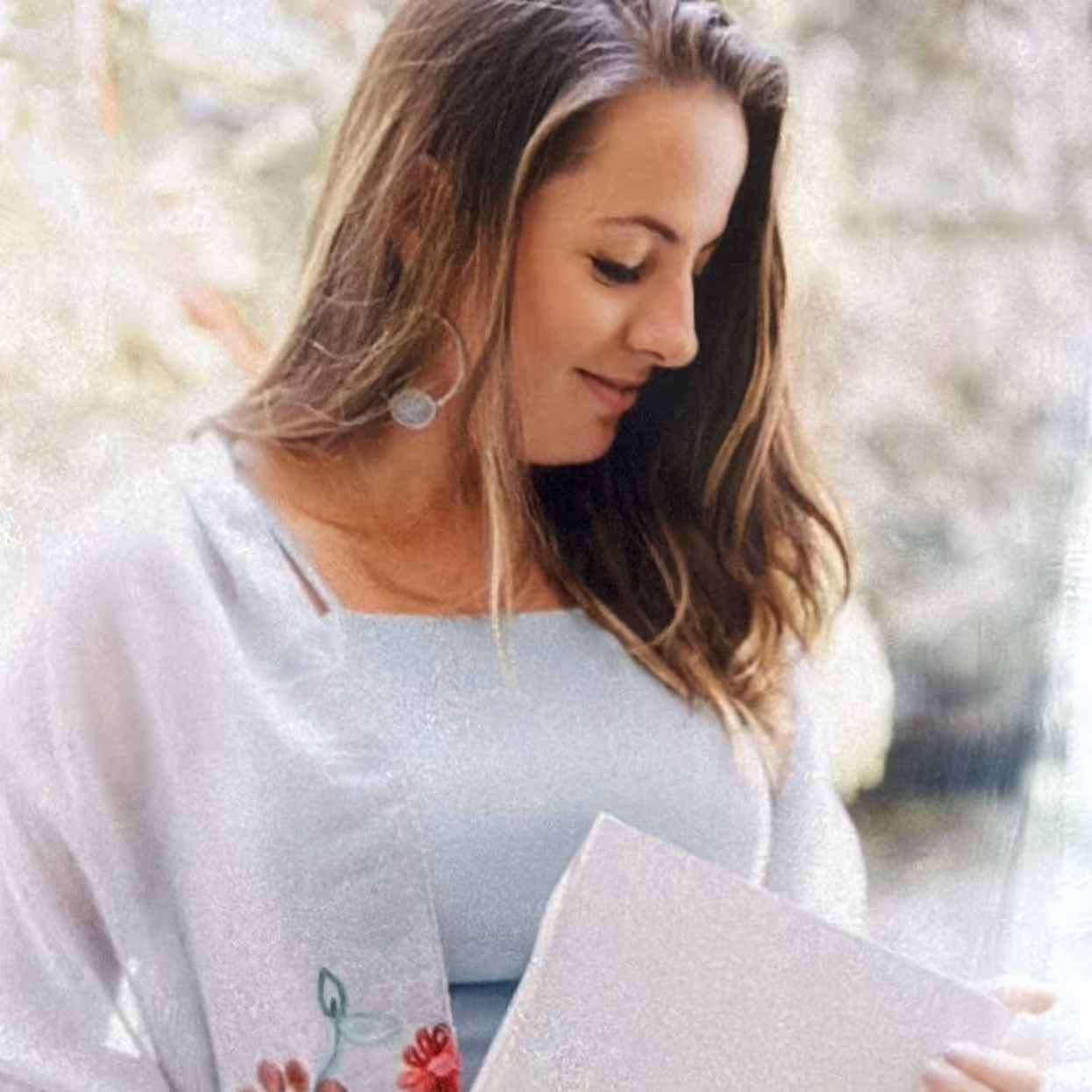 Mulher sorrindo e olhando para baixo segurando um livro ou caderno, com cabelo castanho solto e usando blusa clara com flores bordadas.