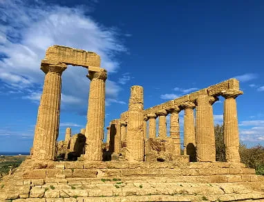 Ruins of an ancient Greek temple with tall stone columns against a blue sky with scattered clouds.