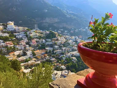 View of a hillside town with numerous white buildings and greenery, with a red flower pot in the foreground.