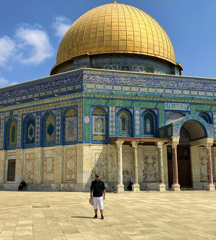 Person standing in front of the Dome of the Rock with its golden dome and intricate blue, green, and gold tilework under a clear blue sky.
