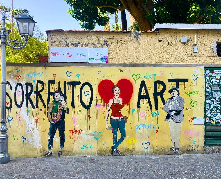 Street mural on a yellow wall featuring the words 'SORRENTO ART' with painted figures of a man, a woman with red heart wings, and another man, surrounded by colorful hearts and graffiti.