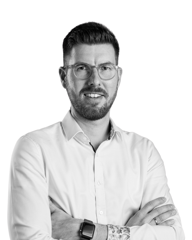 Porträt eines Mannes mit Brille, Kurzhaarschnitt und Bart, der ein weißes Hemd trägt und die Arme verschränkt.