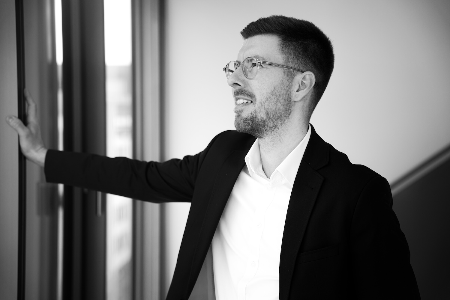 Geschäftsmann mit Brille und Anzug lehnt an Fensterscheibe und blickt nach rechts.