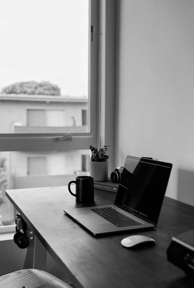Schwarz-weiß-Foto eines Schreibtischs mit einem offenen Laptop, einer Tasse, einer Pflanze, einer Kamera und einer Maus neben einem Fenster.