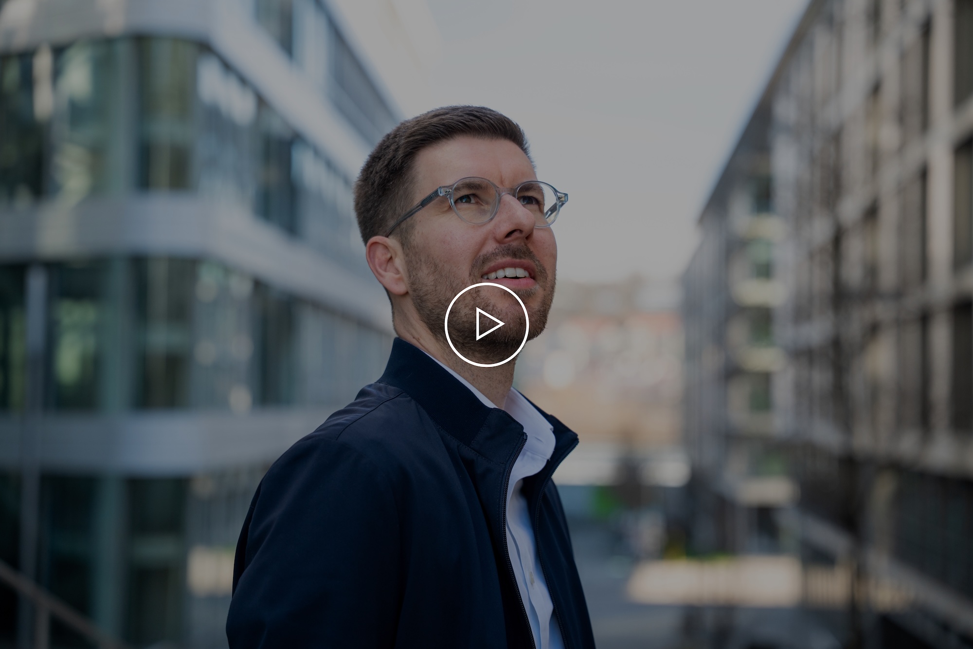 Mann mit Brille und dunkler Jacke blickt nach oben zwischen modernen Bürogebäuden