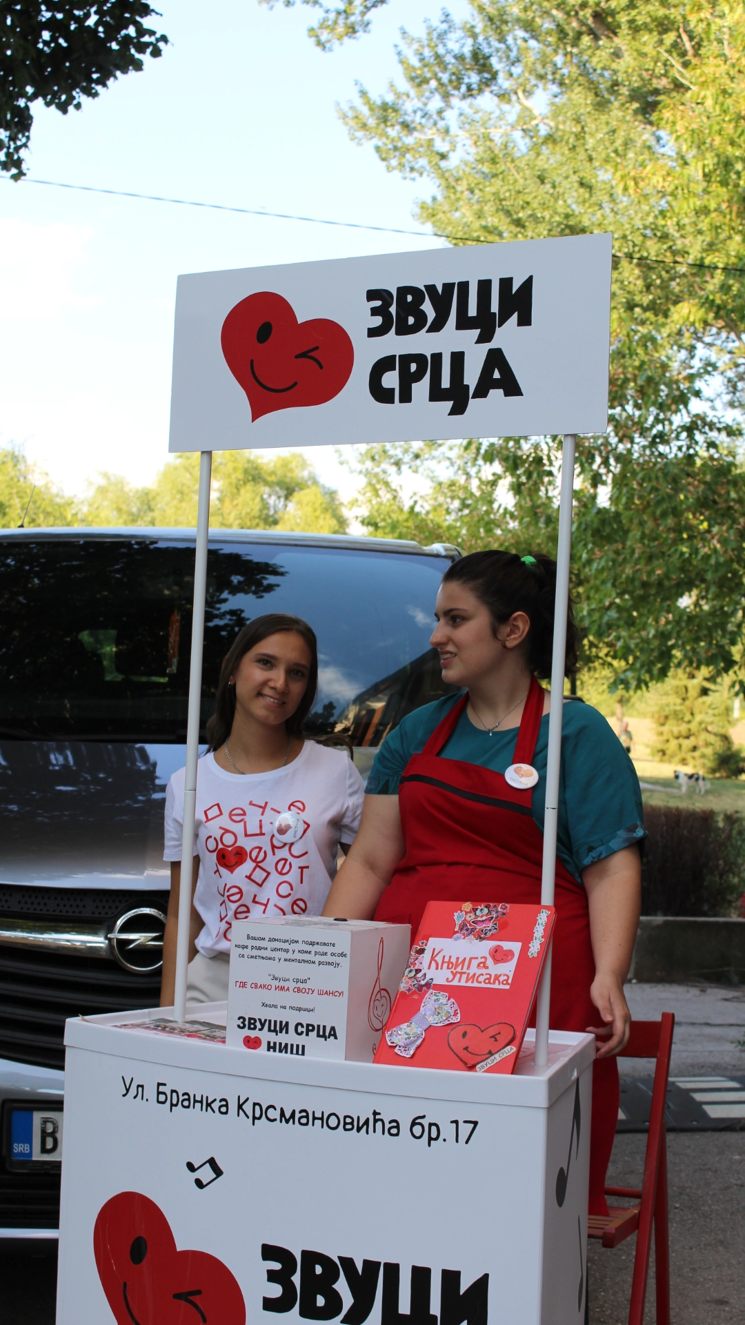 Dve žene stoje iza štanda za donacije sa natpisom Zvuči srca na srpskom jeziku, u obliku srca koje trepće, na otvorenom ispred parka i automobila.