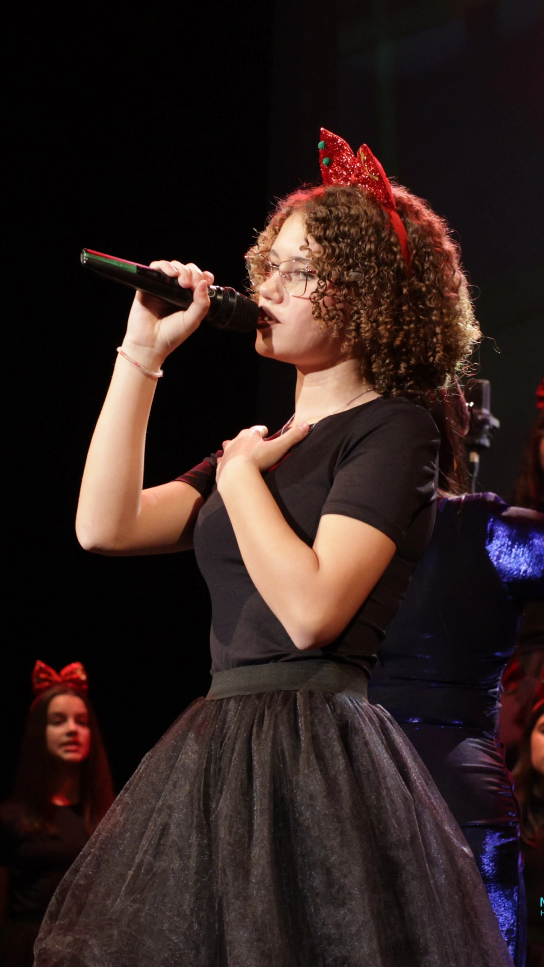Devojka sa kovrdžavom kosom i crvenom trakom sa svetlucavim ukrasom peva držeći mikrofon dok je na sceni.