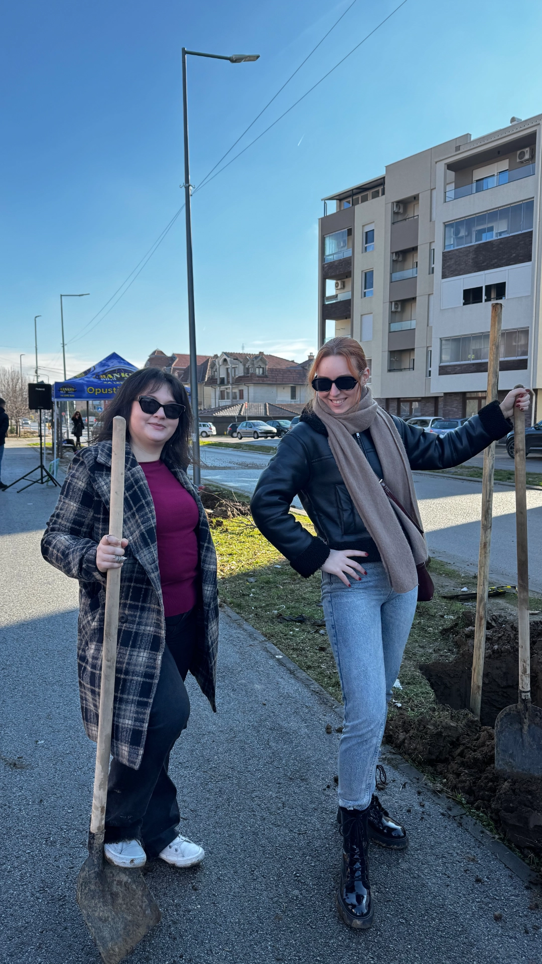 Dve žene sa naočarima za sunce, drže lopate na pločniku pored iskopanih rupa i oblačne su u zimsku odeću.