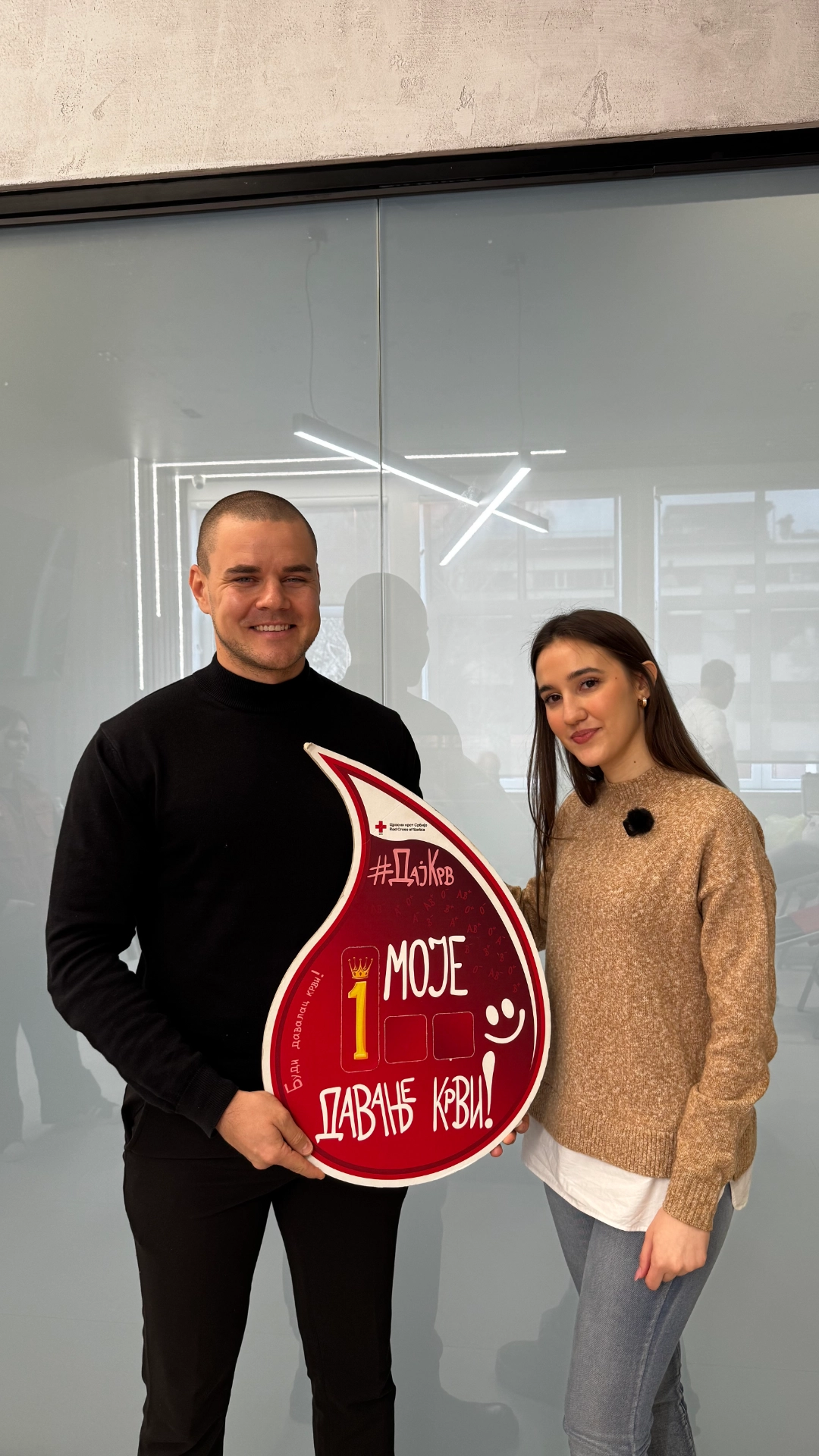 Muškarac i žena drže crveni transparent u obliku kapljice krvi sa natpisom "Moje 1. davanje krvi" u kancelariji.