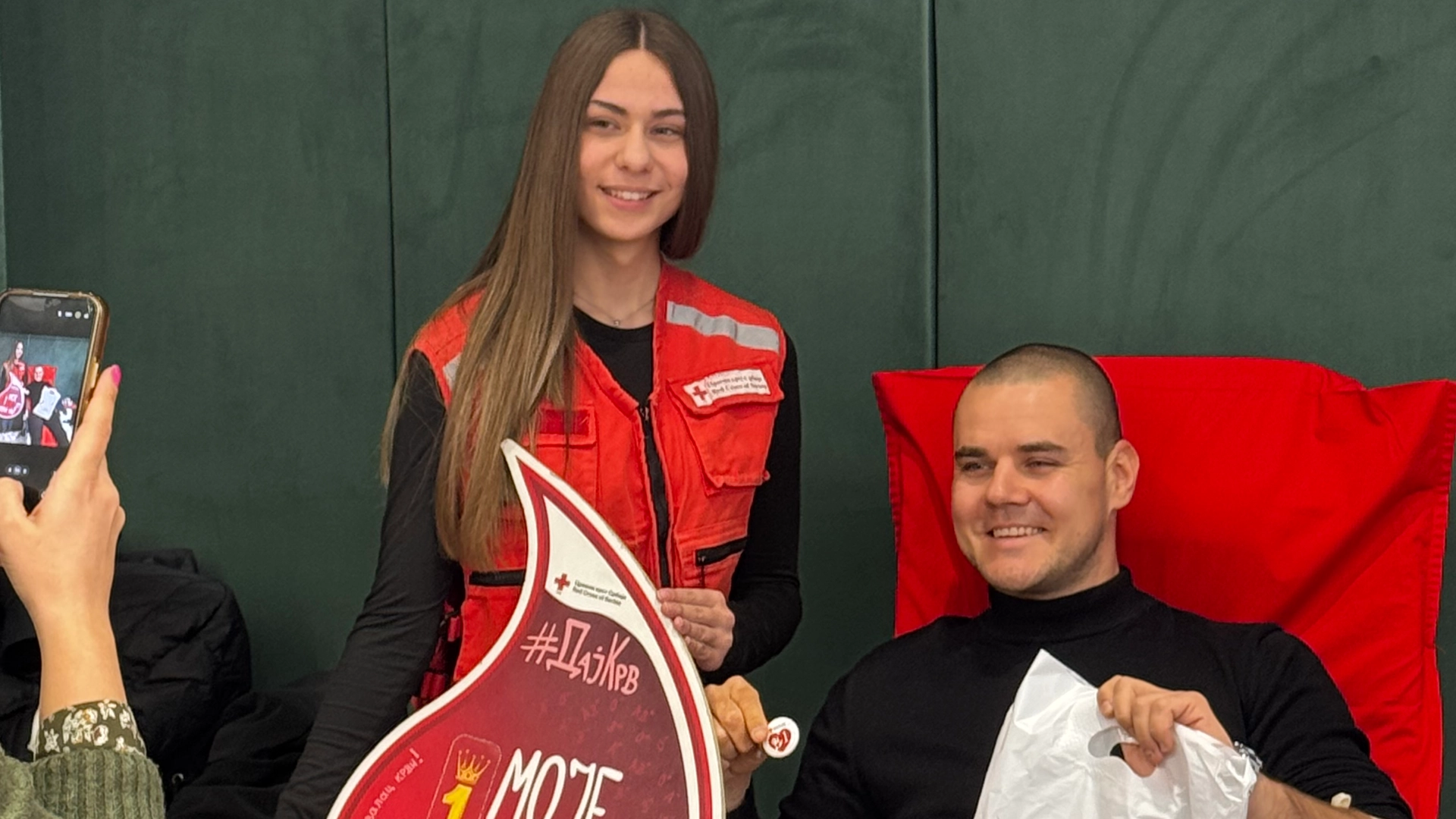 Mlada žena u crvenom prsluku Crvenog krsta stoji pored muškarca koji sedi u stolici za davanje krvi, oboje se smeše.