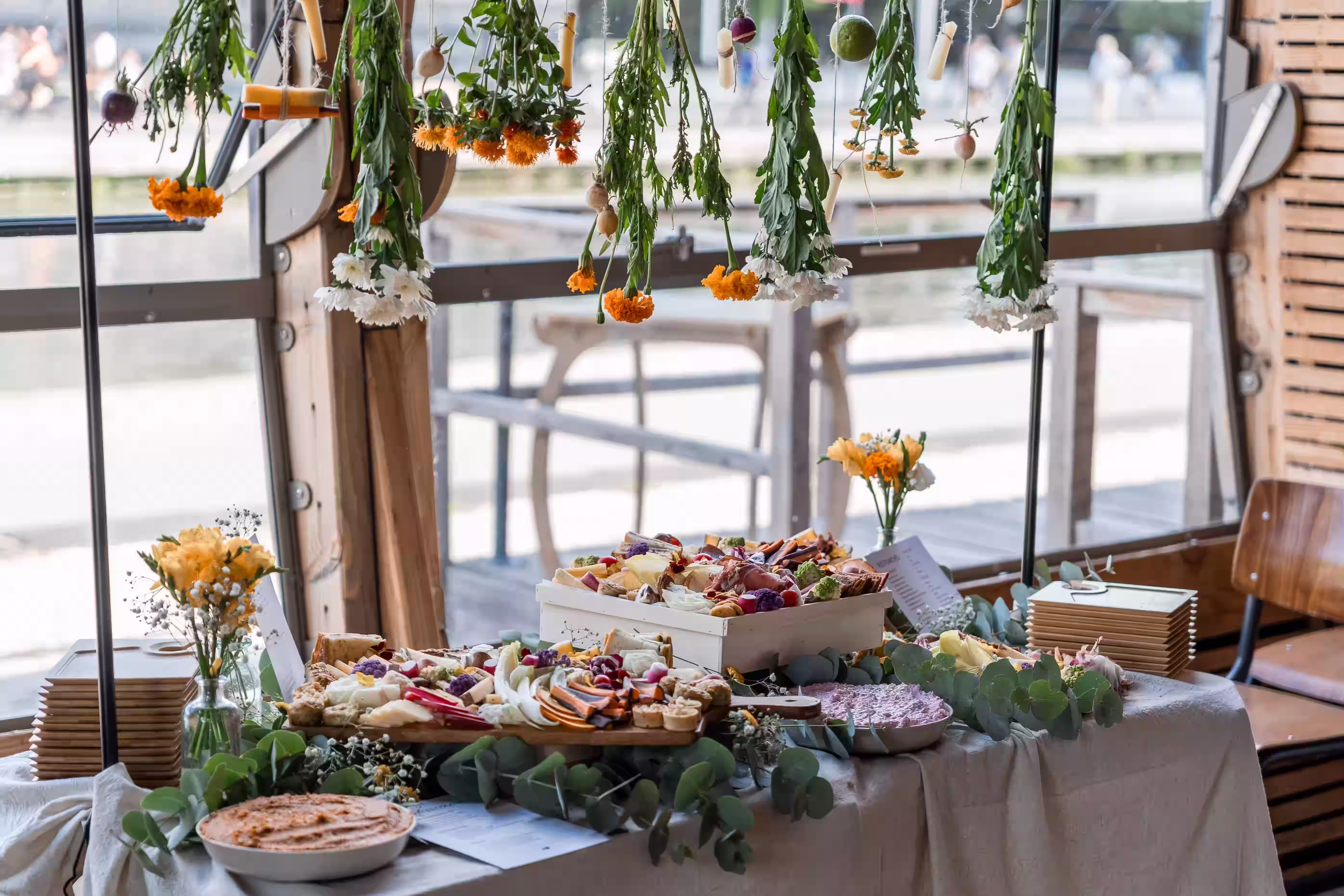 Table de buffet événementiel élégamment décorée avec des compositions florales suspendues, du feuillage d'eucalyptus, entourant des planches en bois garnies de mets colorés et de tartinables.