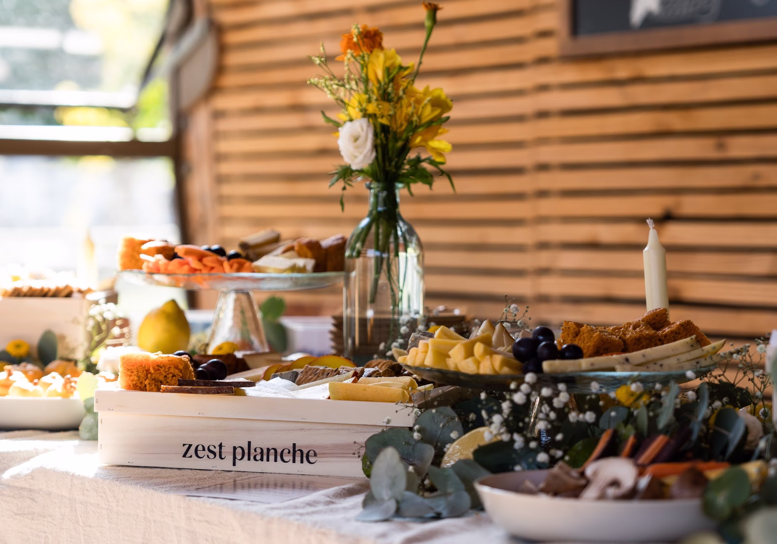 Buffet traiteur décoré avec soin grâce à notre service de scénographie florale : un bouquet de fleurs fraîches trône au milieu d'une planche en bois Zest Planche garnie de fromages.