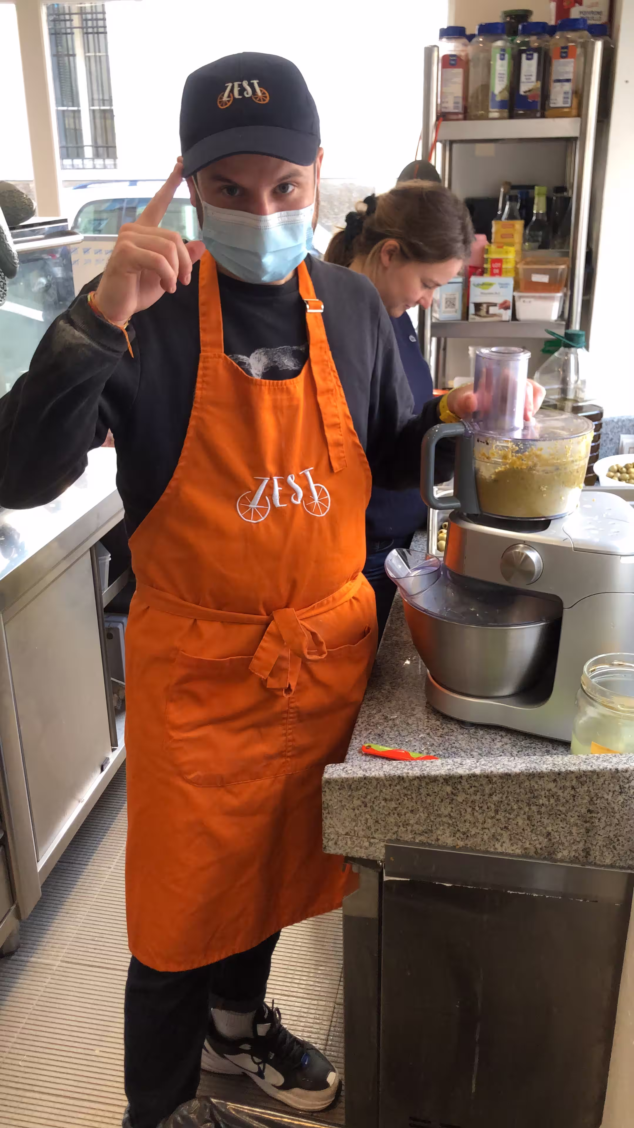 Un cuisinier de l'équipe Zest Planche, portant un tablier orange au logo de la marque, prépare une recette maison à l'aide d'un robot culinaire dans l'atelier parisien.