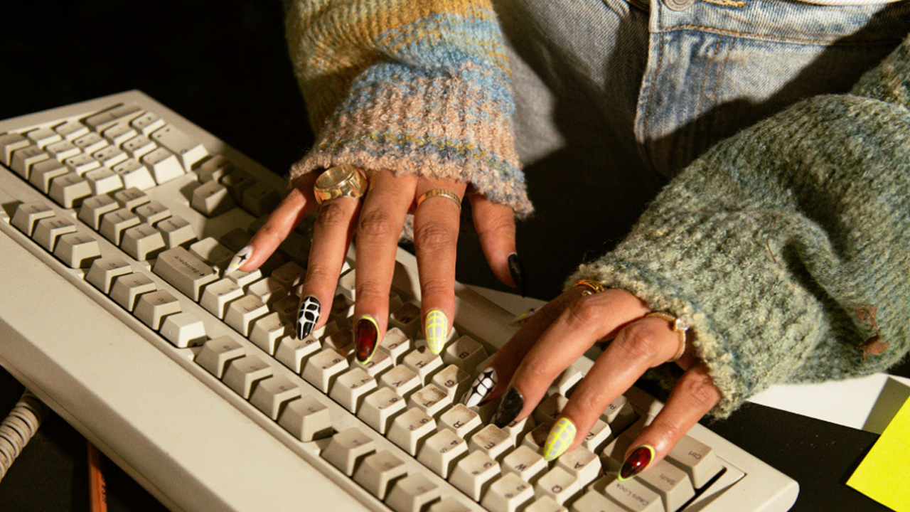 Close-up of hands with nail art and gold rings typing on a vintage beige keyboard