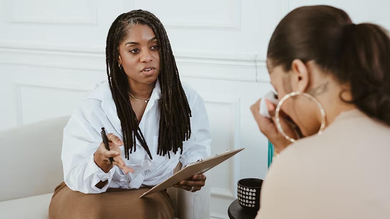 Therapist holding a clipboard and pen talking with a client who's wiping her eyes with a tissue
