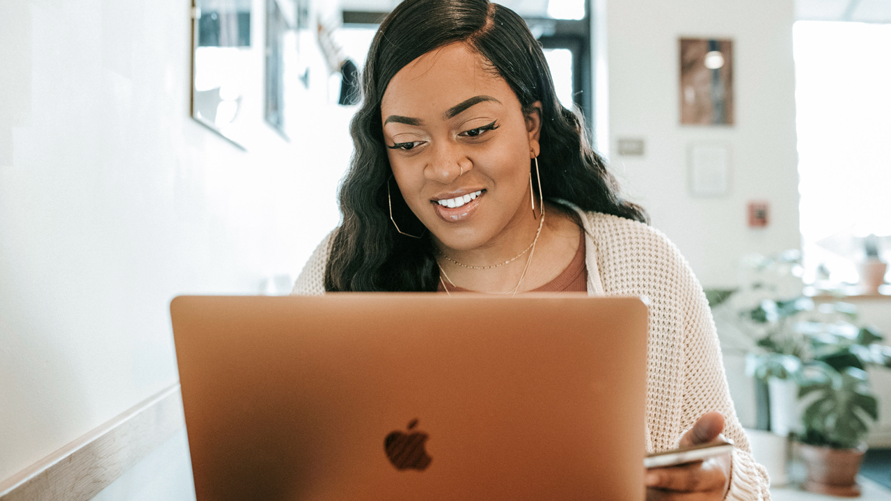 Pittsburgh website designer Domanique Taylor in hoop earrings and a cream cardigan smiling at her laptop screen
