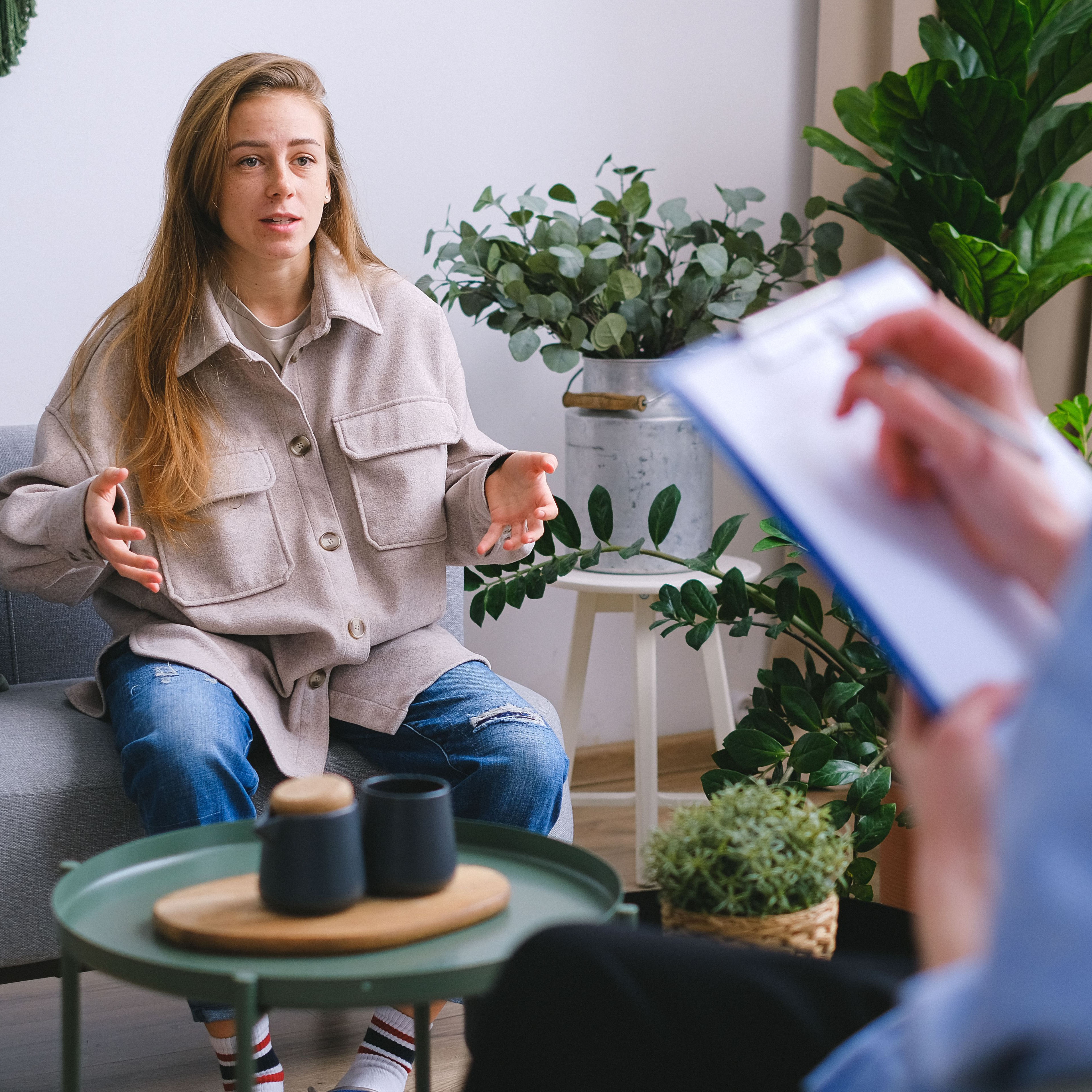 Woman in a tan jacket talking with a therapist who is taking notes, in a room filled with plants