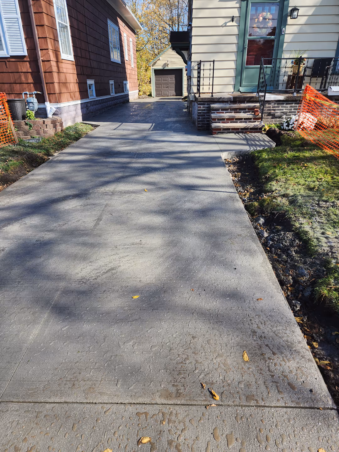 Newly poured concrete driveway leading to a garage behind two houses, with orange safety fencing on both sides.