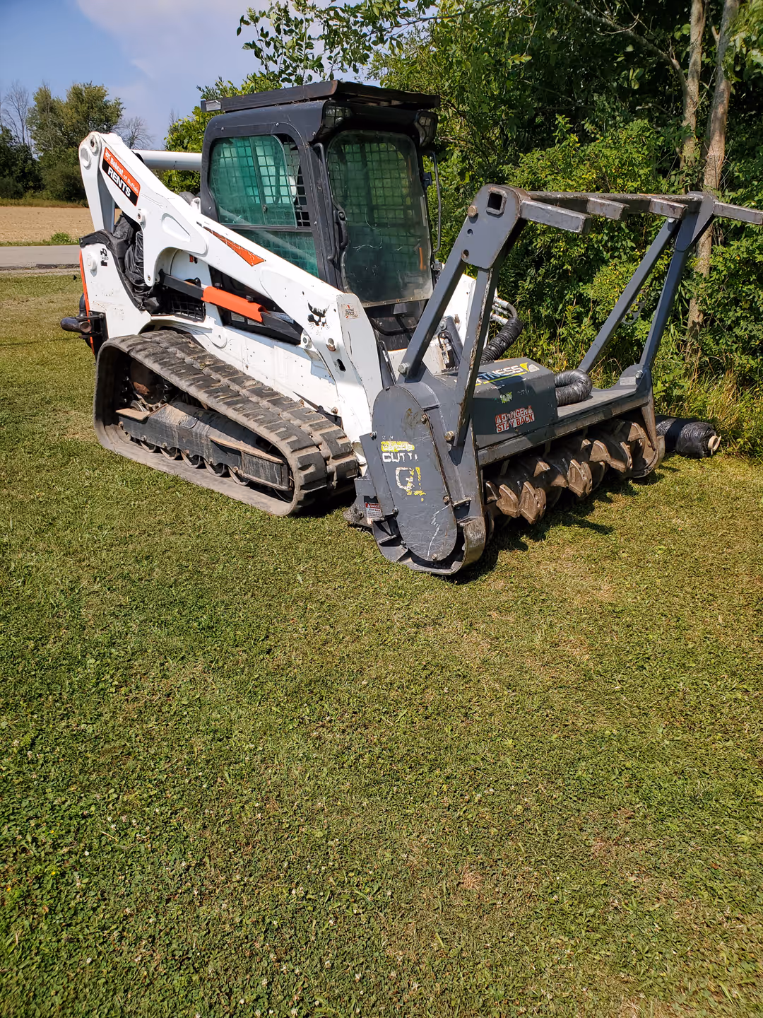 White compact track loader with a large soil tiller attachment parked on grass near trees.