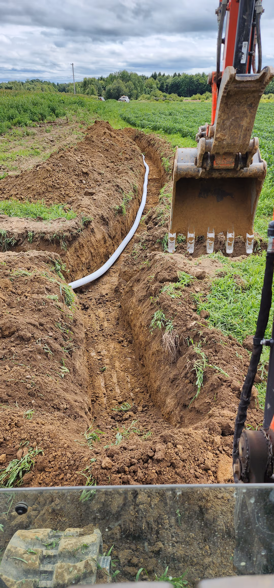 Excavator digging a trench in a green field with a white pipe placed inside the trench under a cloudy sky.