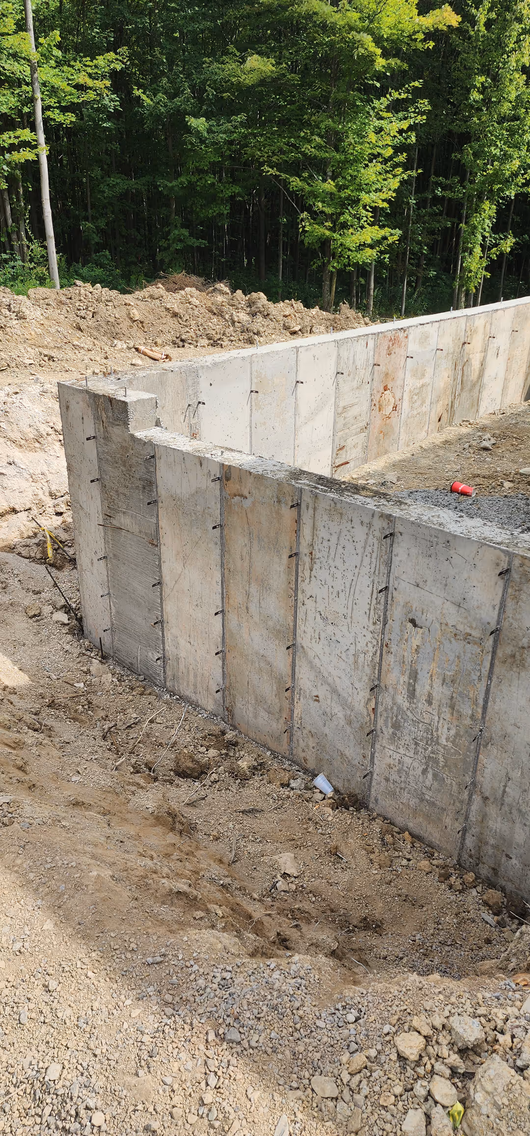 Concrete foundation wall under construction with exposed dirt and a forest in the background.
