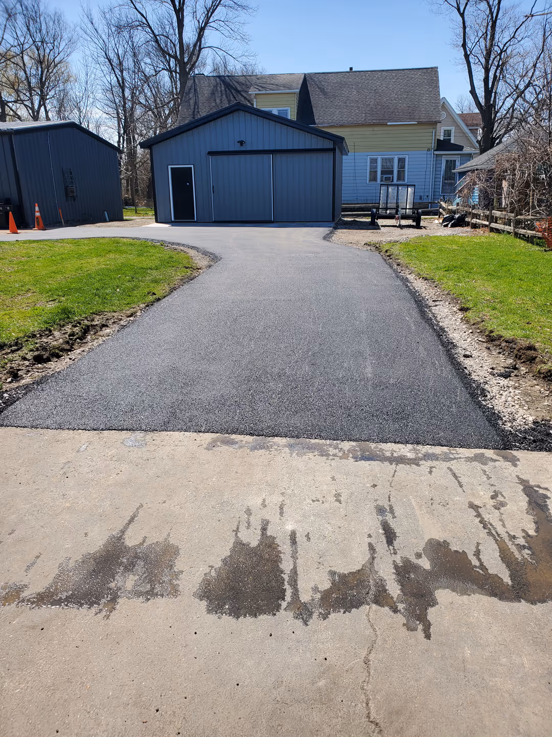 Newly paved black asphalt driveway leading to a gray garage with a black door, flanked by green lawns and leafless trees on a clear day.